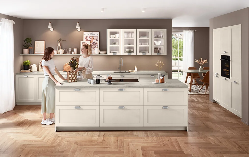 Moderne Landhausküche in Sand matt mit Front Nordic 788, Kochinsel, klarer Linienführung und heller Travertin-Arbeitsplatte vor warmem Wandton.