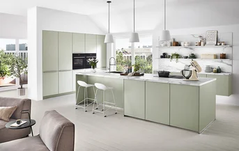 Helle grifflose Küche mit Front SENSO 494 in Jade Premium matt, Kochinsel mit Marmoroptik-Arbeitsplatte und offenen Regalen vor großer Fensterfront im modernen Wohnraum.