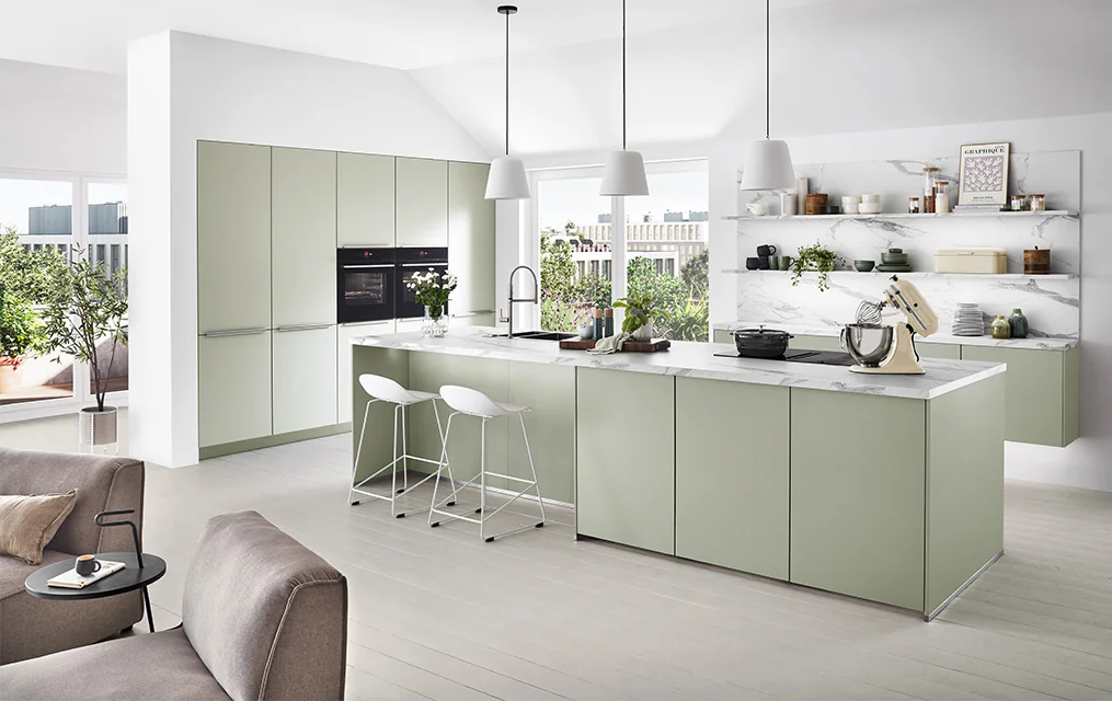Helle grifflose Küche mit Front SENSO 494 in Jade Premium matt, Kochinsel mit Marmoroptik-Arbeitsplatte und offenen Regalen vor großer Fensterfront im modernen Wohnraum.