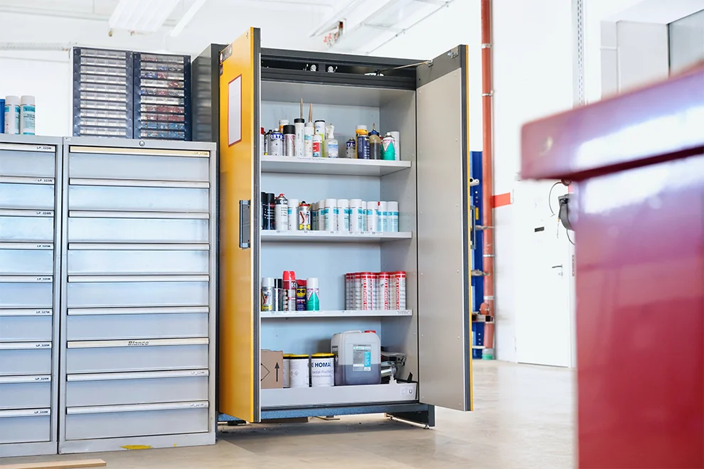 Un atelier bien organisé avec une grande armoire de rangement ouverte affichant diverses boîtes, bouteilles et équipements, à côté d'une unité de tiroirs en métal.