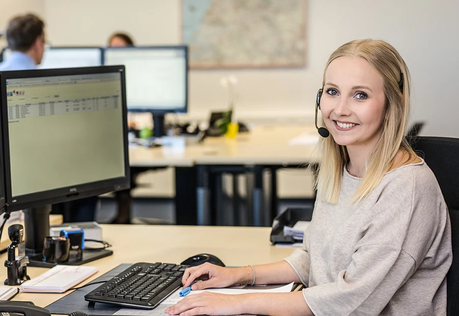 Professioneller Kundendienstmitarbeiter mit Headset lächelt in die Kamera in einem modernen Büro, was eine einladende Unternehmensumgebung zeigt.
