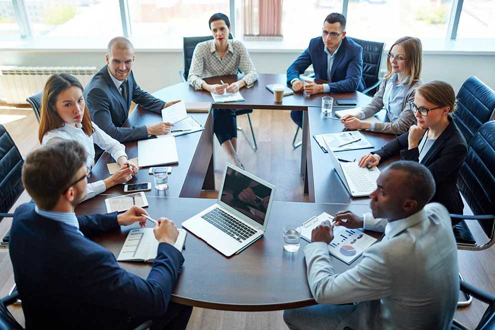 Groupe diversifié de professionnels engagés dans une réunion autour d'une table avec des ordinateurs portables, des documents, et discutant des stratégies commerciales dans un bureau moderne.