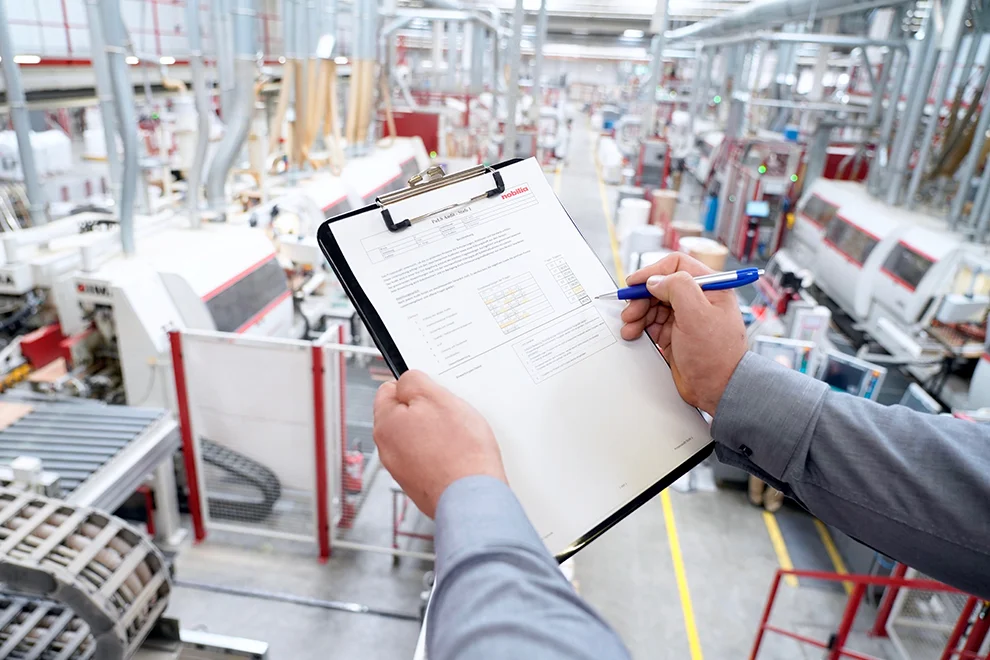 Vue de dessus de l'épaule d'une personne inspectant une liste de contrôle sur un presse-papiers dans une usine de fabrication industrielle moderne avec des machines en arrière-plan.