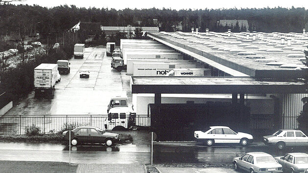 Luftaufnahme des nobilia Werks mit Lkw und Pkw auf nasser Fahrbahn vor der modernen nobilia Gebäudeansicht im Jahr 1985.